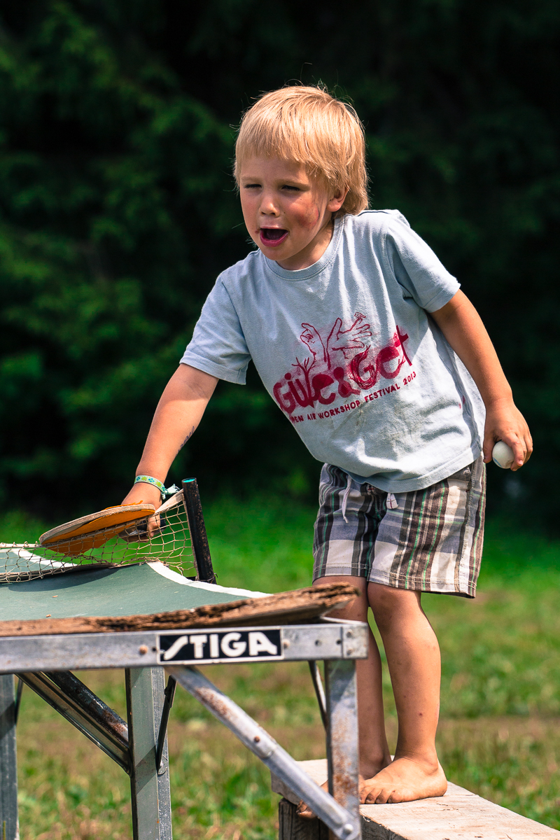 A young blond boy is standing on a wooden platform by a table tennis table at the 2013 Give and Get Festival. He is holding a table tennis paddle in one hand and a ball in the other. The boy is wearing a light blue T-shirt with red graphics and plaid shorts. The background shows greenery, suggesting an outdoor setting.