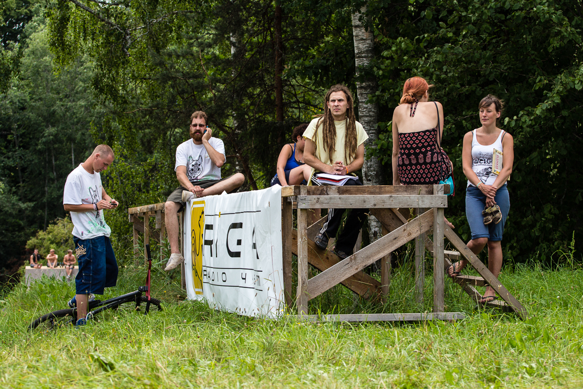 A group of people gather around a wooden platform in an outdoor setting surrounded by trees during the 2013 Give and Get Festival. Some are sitting on the platform, while others stand nearby. One person is writing in a notebook. There is a banner with text and a logo partially visible on the platform's side.