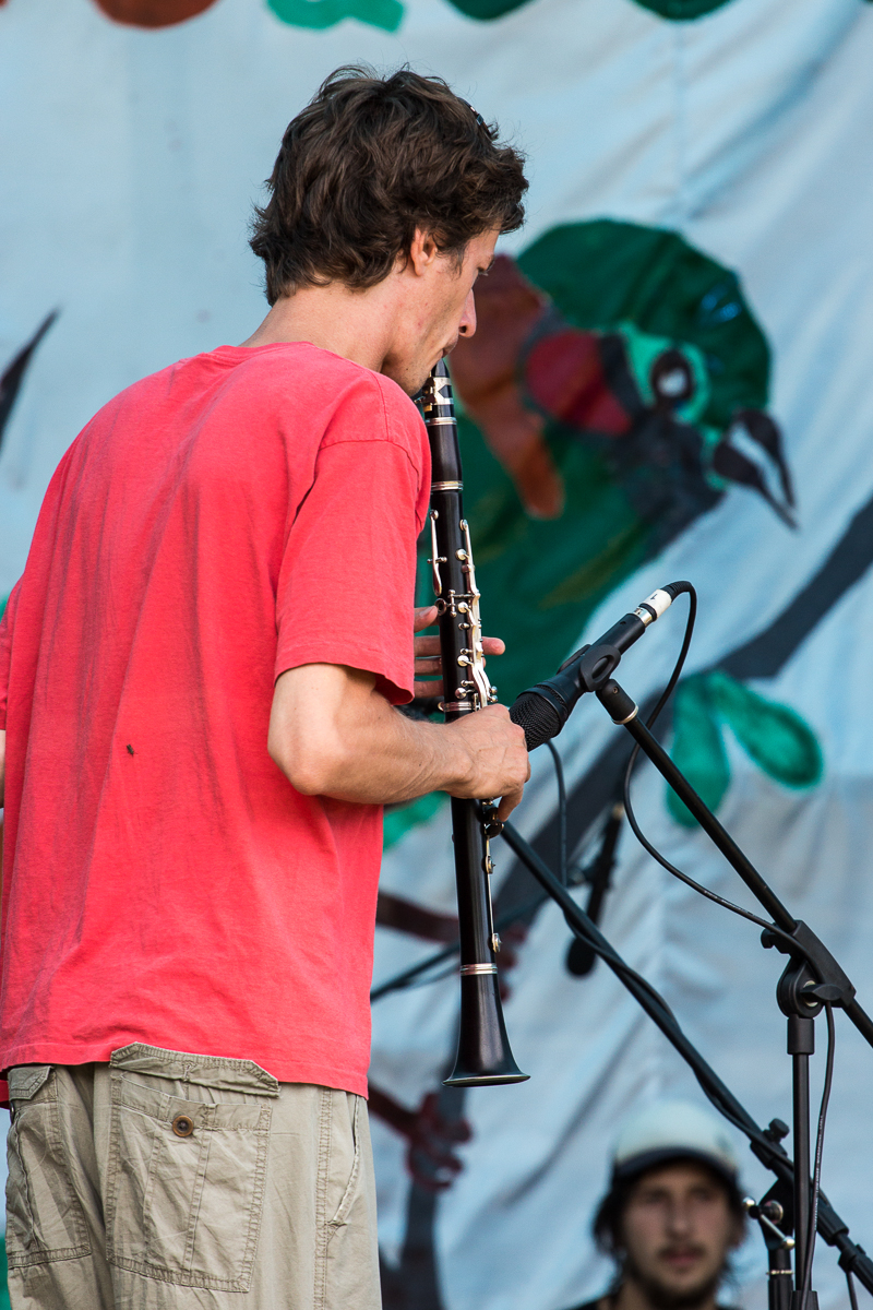 A man in a red T-shirt and khaki shorts plays a clarinet on stage at the 2013 Give and Get festival, facing away from the camera. Behind him is a microphone stand. Another person in a cap is blurred in the background. The backdrop features colorful illustrations of birds and greenery.