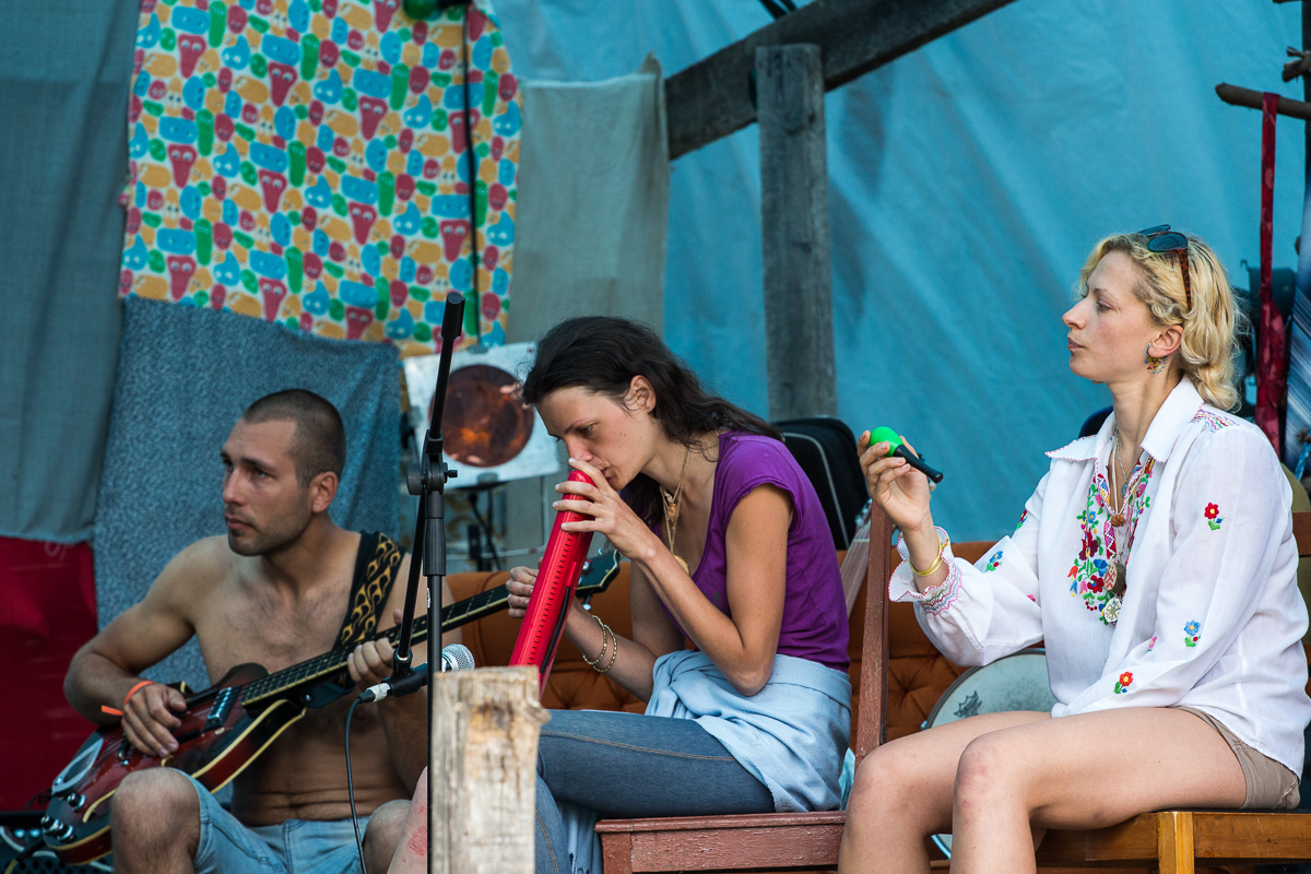 Three people sitting on stage appear to be preparing for a musical performance at the 2013 Give and Get festival. The man on the left, shirtless, holds a guitar. The woman in the center blows into a melodica, while the woman on the right, wearing a white embroidered top, looks ahead, holding a maraca. The background is colorful and eclectic.