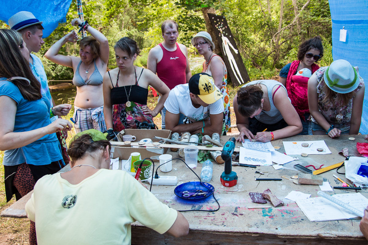 At the 2013 Give and Get festival, a group of people gather around a table outdoors under a blue tarp. Some are standing, while others are seated, focusing on various craft activities. The table is cluttered with art and craft materials like papers, tools, and rocks. Trees and greenery provide a scenic backdrop.