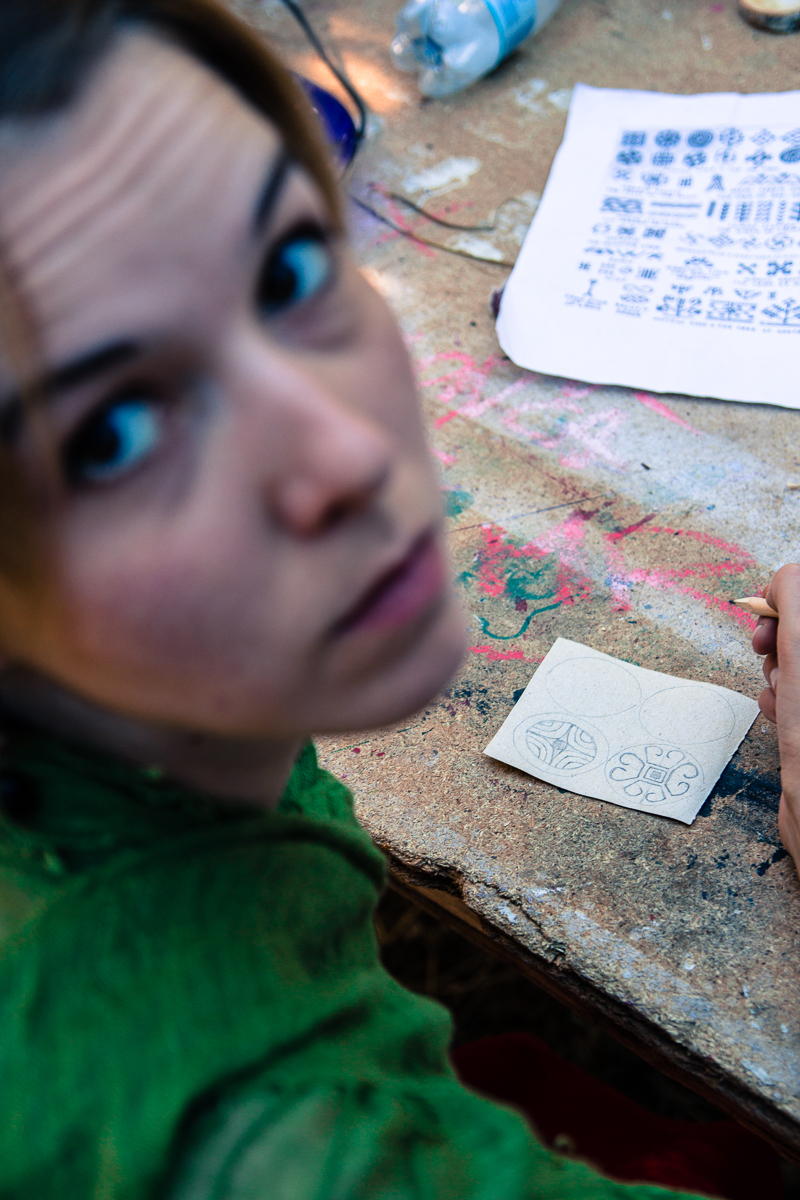 A woman with light-colored hair, wearing a green top, sits at a cluttered table looking up towards the camera. She holds a pen and is drawing intricate patterns on a small piece of paper. Nearby on the table is another paper with similar pattern sketches and various art supplies scattered around, reminiscent of the creative ambiance at the 2013 Give and Get festival.