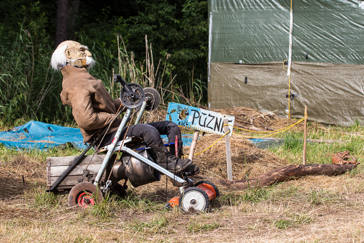 A scarecrow dressed in a brown jacket, black pants, and a worn mask with white hair sits on a makeshift vehicle made from scrap materials and an old lawnmower. Behind it, a hand-painted sign reads "PUZN," reminiscent of the 2013 Give and Get festival, surrounded by greenery and a tarp-covered structure. The ground is covered in dry grass.