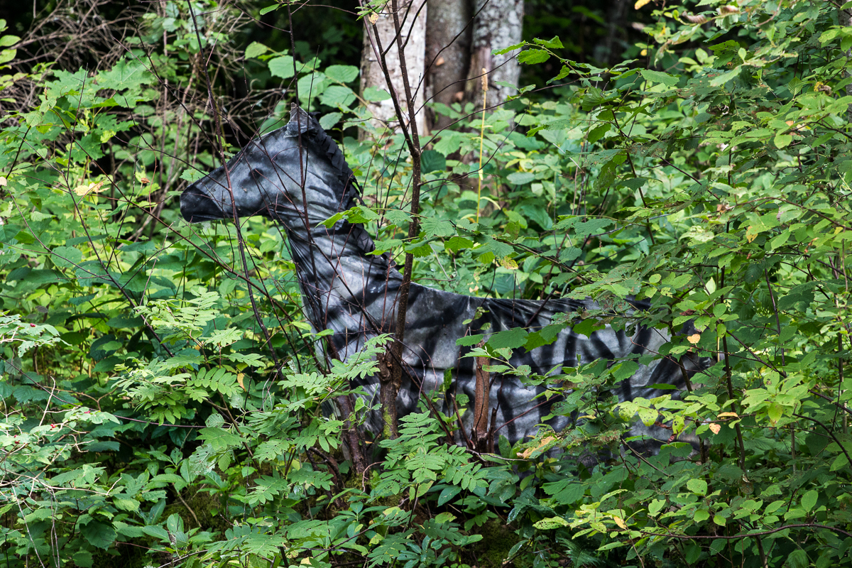 A metallic deer statue, partially concealed by lush green foliage, stands amidst a dense forest. Sunlight filters through the trees, casting dappled shadows on the statue and surrounding leaves, creating a blend of light and dark patches throughout the scene—an artwork from the 2013 Give and Get festival.
