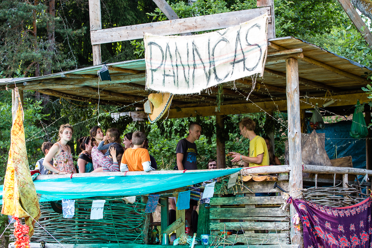 A group of people stands and sits under a wooden, open-sided shelter in a forested area during the 2013 Give and Get festival. A large hand-painted sign that reads "PANNCAKES" hangs at the front. Strings of lights and colorful fabrics decorate the space, giving it a festive look. Trees and greenery are visible in the background.