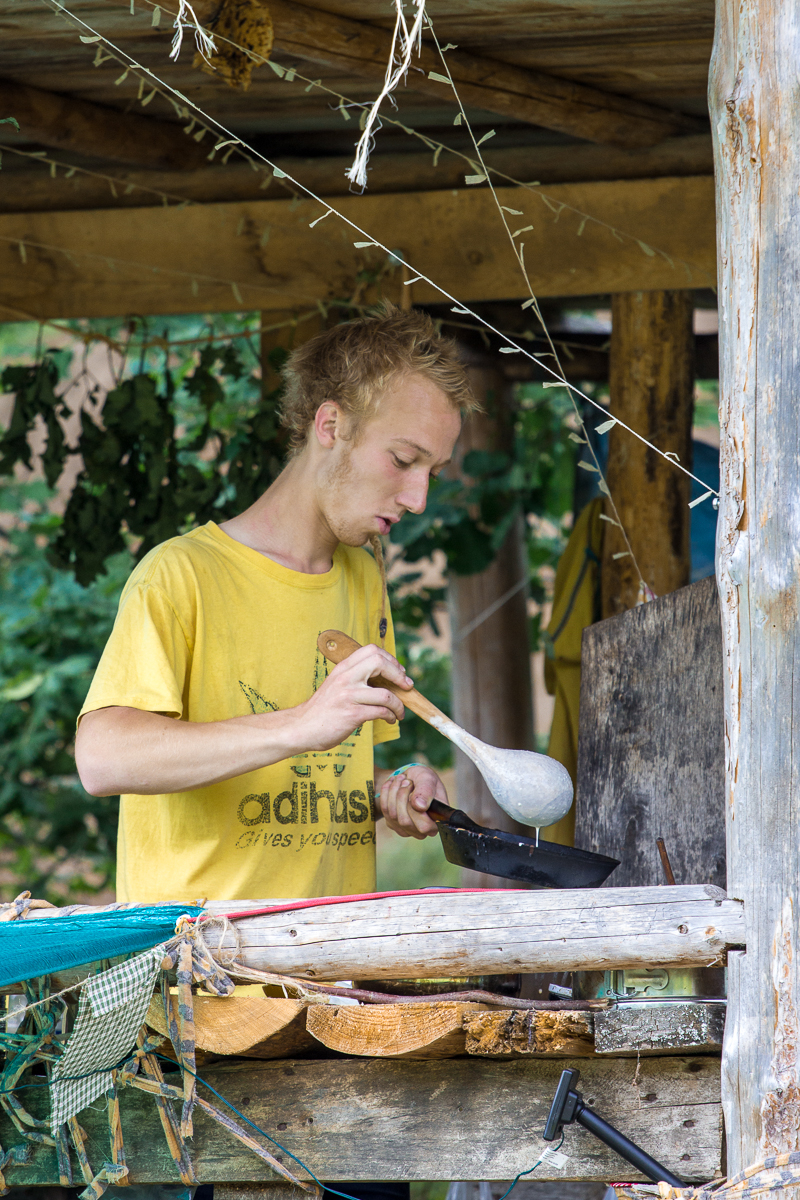 A young man with blond hair is under a rustic wooden shelter, wearing a yellow t-shirt with an illustration from the Give and Get Festival 2013. He is holding a large wooden spoon and appears to be cooking in a black frying pan. Various kitchen items and greenery surround the rustic setting.