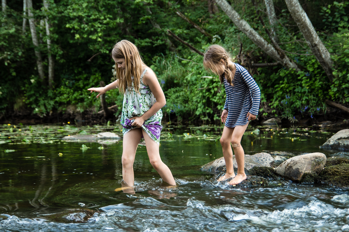 Two young girls are seen wading through a shallow, rocky stream at the Give and Get festival. The girl on the left wears a multicolored sleeveless dress, while the girl on the right sports a blue-and-white striped dress. Both are barefoot, carefully stepping on rocks amidst lush green trees and vegetation.