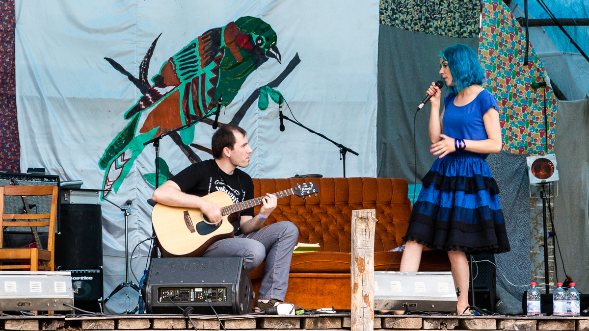 A man seated playing an acoustic guitar and a woman with blue hair standing with a microphone perform on an outdoor stage at the 2013 Give and Get festival. Behind them is a colorful bird mural and patterned fabric. An orange-brown couch and various musical equipment, including speakers, complete the scene.
