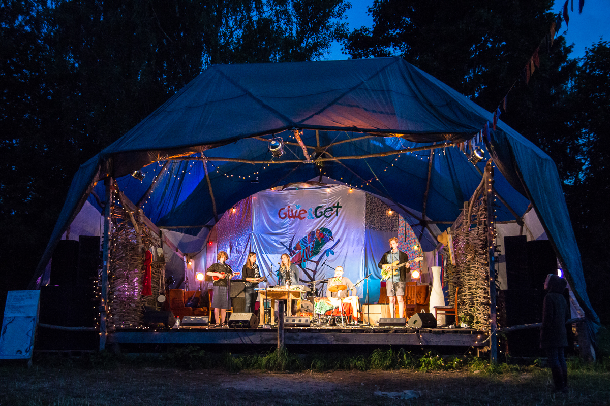 A band performs on an outdoor stage at night during the 2013 Give and Get festival. The stage, adorned with blue and white drapes and lights, hosts musicians playing drums, guitar, and keyboard. Trees frame the scene while an audience member watches from the right.