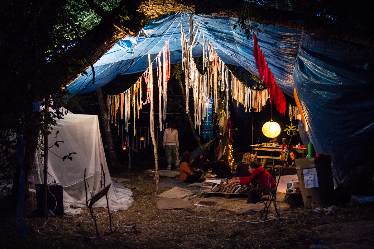 A cozy outdoor setup under a large blue tarp is visible, with various hanging white and red fabric decorations. A group of people are sitting on the ground around a lantern, engaging in activities reminiscent of the Give and Get festival. A white tent-like structure is on the left, and the surrounding area is dimly lit with a warm glow.