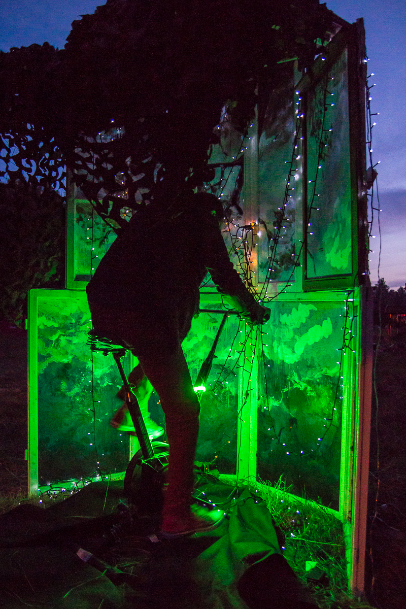 A silhouetted person operates an illuminated art installation at the 2013 Give and Get festival. The scene is lit with vibrant green lights, with string lights draped along the frame and surrounding structure. The dark background highlights the glowing elements of the installation beautifully.