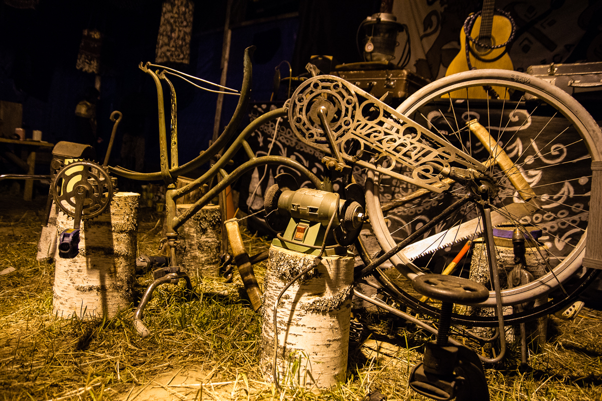A cluttered, rustic workshop with various tools and metal parts. In the foreground, a stationary bicycle wheel is mounted on a log. Nearby, an old grinder and other metalworking tools are placed on tree stumps. The background shows an old guitar and various hanging items from the Give and Get festival 2013 in a dimly lit space.