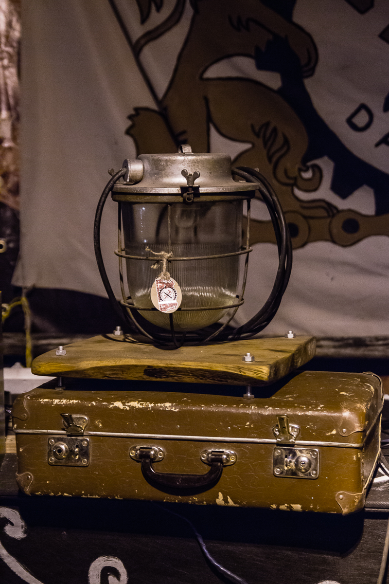 An antique miner's lamp with a glass encasement and metal top is displayed atop a worn, brown leather suitcase. The lamp has a small emblem attached to it. The background features a partially visible banner with a stylized lion design from the 2013 Give and Get festival. The scene is dimly lit.