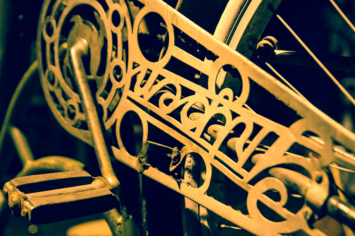 Close-up view of a vintage bicycle crank and chain guard with intricate geometric cutouts. The metal parts have a weathered, golden patina, reminiscent of the decor at the 2013 Give and Get event. The image features an artistic, slightly faded look, highlighting the detailed craftsmanship of the old bicycle components.