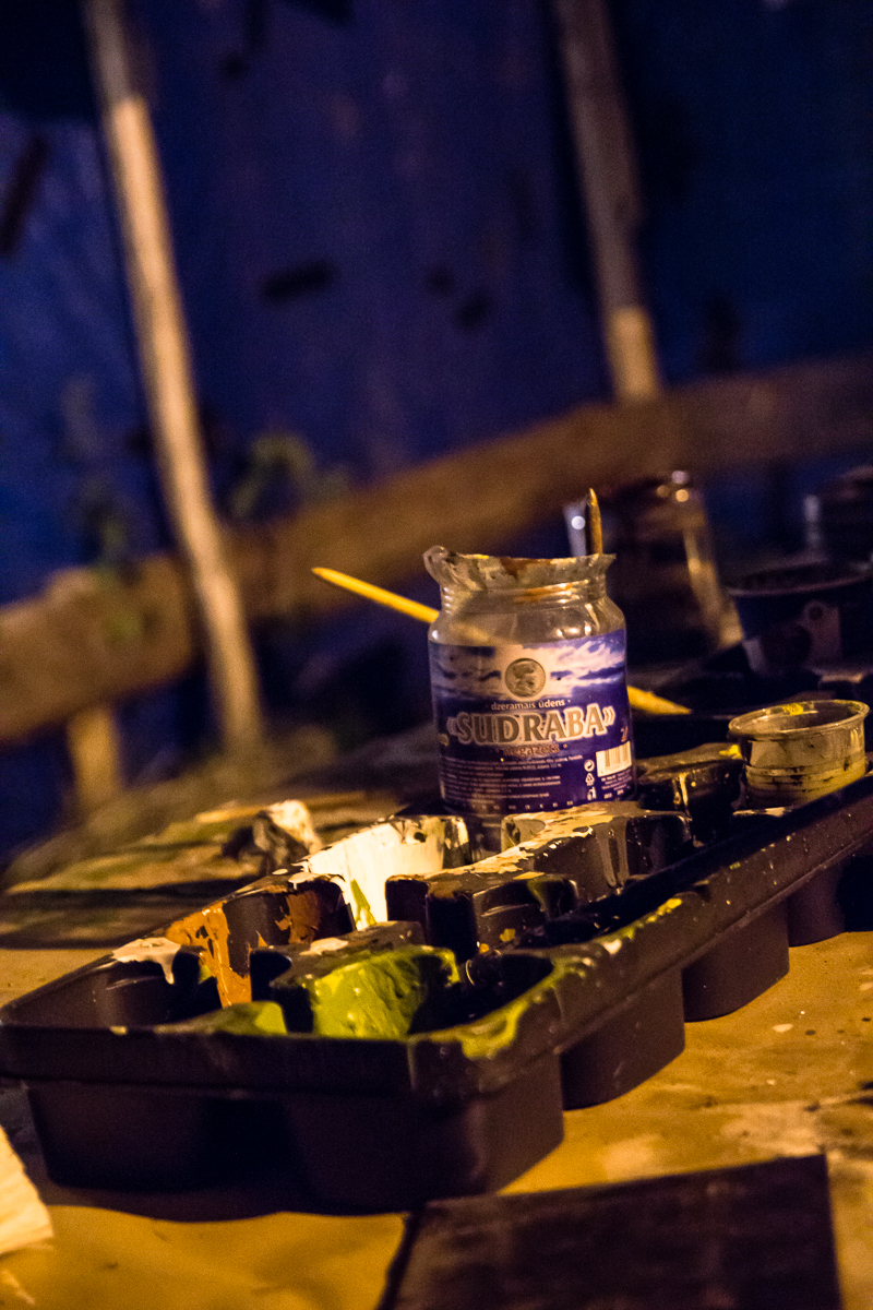 A dimly lit image of a workspace featuring a plastic paint palette with several compartments filled with various shades of paint, remnants from the 2013 Give and Get festival. A partially used paintbrush rests atop the palette. In the background, an open jar labeled "SUDRABA" contains liquid, with a yellow paintbrush jutting out.