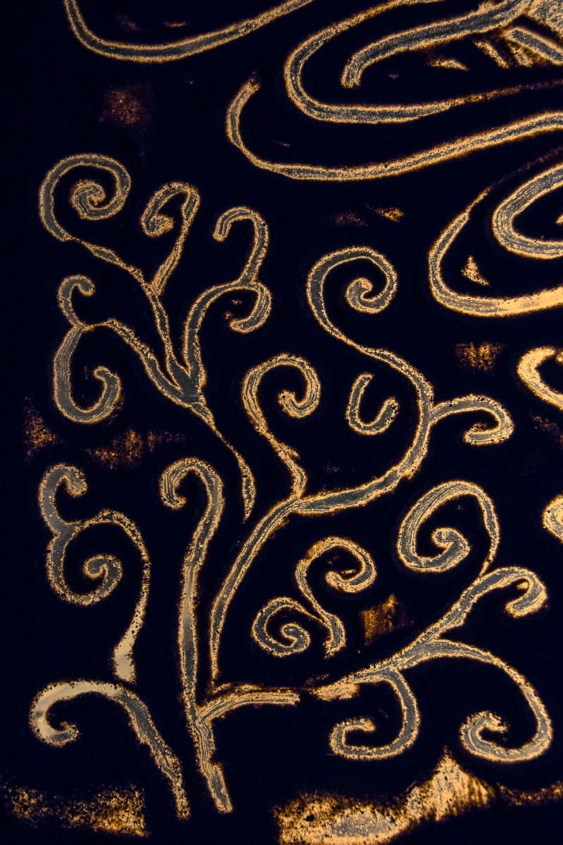 A dark artwork featuring intricate, swirling patterns drawn with a golden hue. The flowing lines form abstract, organic designs reminiscent of vines or tendrils, set against a deep black background, evoking the elegance of the Give and Get festival 2013's vibrant spirit with its striking contrast and decorative appearance.