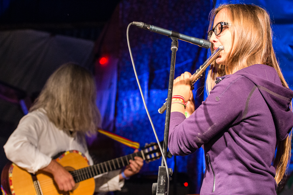 A young woman in glasses and a purple hoodie plays the flute into a microphone on stage at the 2013 Give and Get festival. In the background, an older person with long gray hair plays a guitar. The scene is lit with blue and purple stage lights, suggesting a live musical performance.