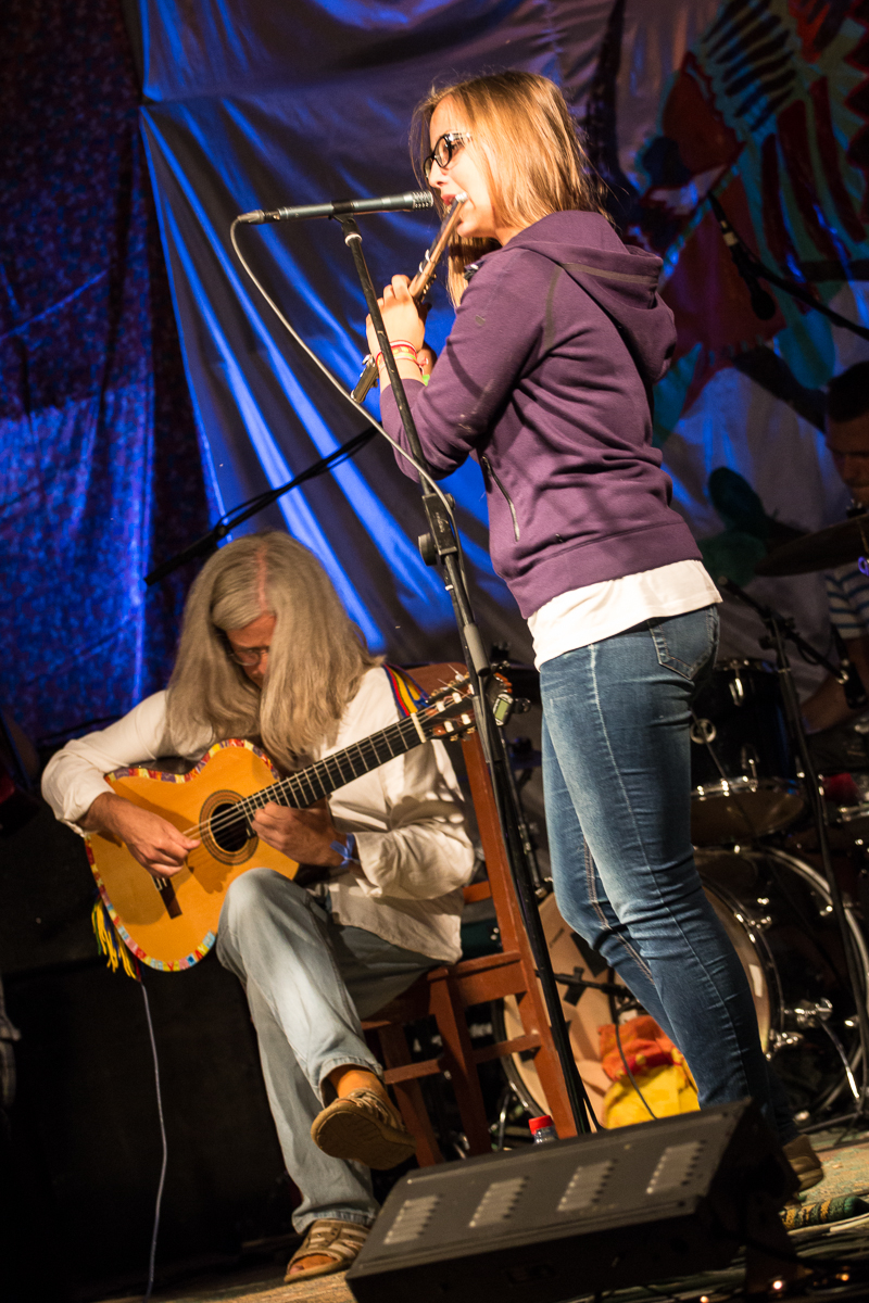 At the Give and Get festival 2013, a woman wearing a purple hoodie and jeans plays a flute into a microphone on stage. Beside her, a man with long white hair in a white shirt and jeans strums a yellow acoustic guitar. The background is decorated with colorful fabric, and a drum set is visible behind them.