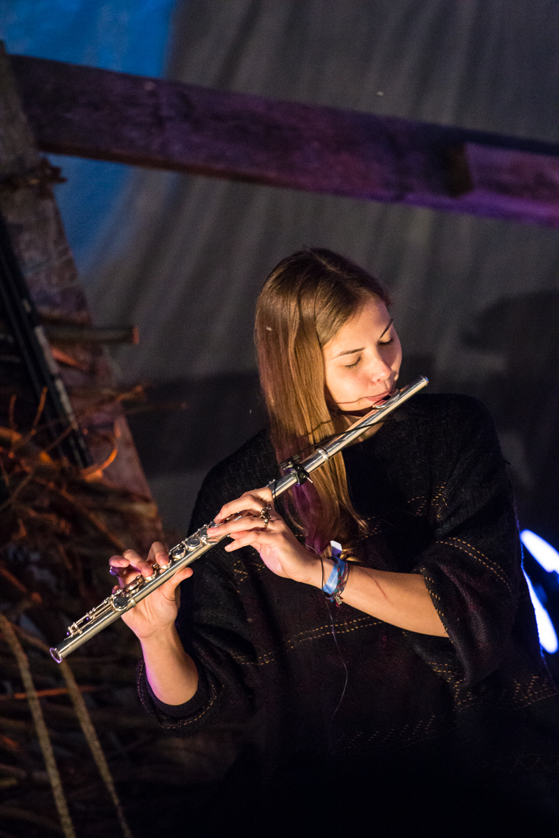 A person with long, light brown hair plays a flute, wearing a black sweater and standing in a dimly lit environment. The background features wooden beams and soft lighting, creating a cozy atmosphere reminiscent of the Give and Get festival. The person appears focused and passionate about their music.