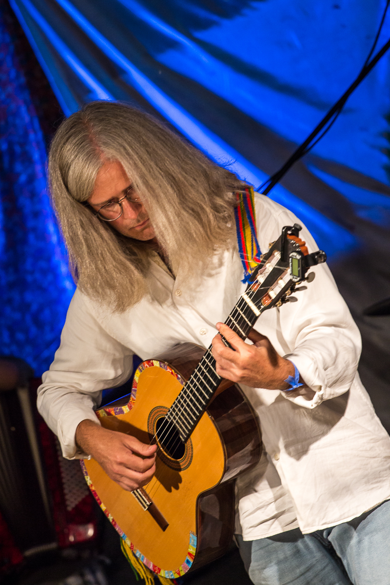At the 2013 festival, a person with long gray hair and glasses plays an acoustic guitar. Dressed in a white shirt and using a multicolored strap, they perform against the blue-lit drapery backdrop, adding colorful ambiance. The musician appears deeply focused and absorbed in the music.