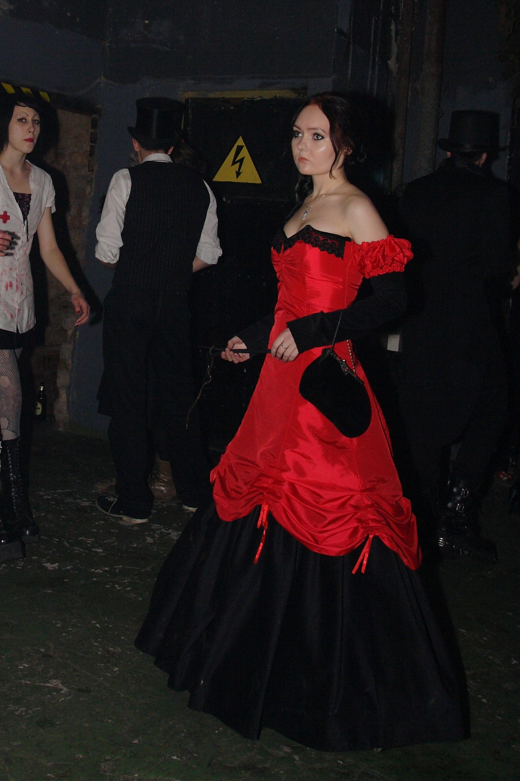 A woman in a vivid red and black Victorian-style gown stands in a dimly lit room, reminiscent of Elektra '09, holding a black fan. She has fair skin and dark hair styled in an updo. Other people in the background wear dark, gothic attire, including top hats and suits, setting the scene for an eerie Halloween party amidst exposed brick and caution signs.