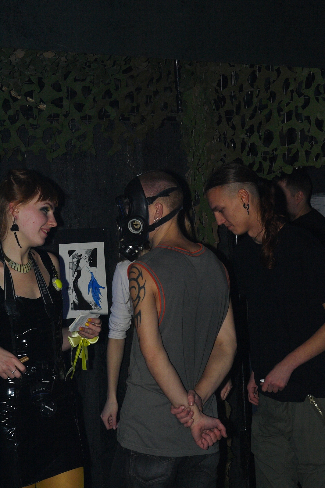A group of four people are seen interacting in a dimly lit room with dark walls and camouflage netting in the background, reminiscent of an Elektra '09 Halloween party. One person wears a sleeveless shirt and a gas mask, while another has long hair pulled back and wears a black shirt. A woman dressed in a shiny black outfit holds a framed picture.
