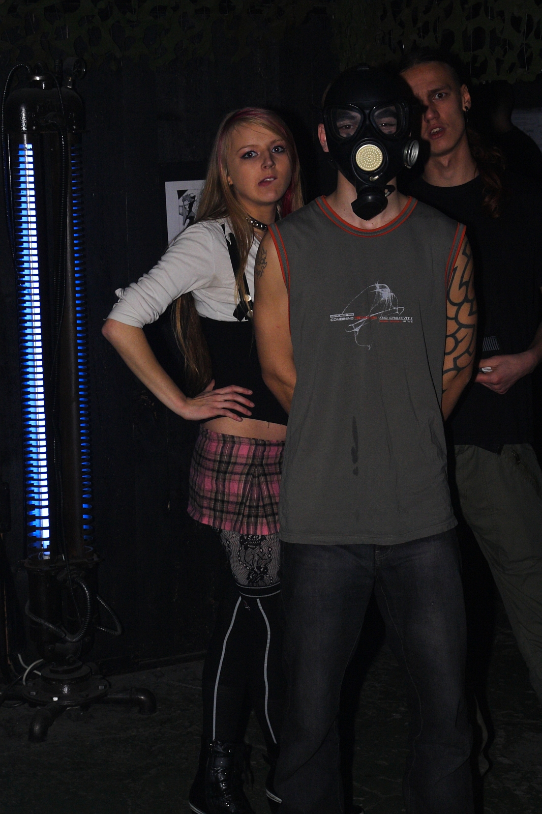 Three people, one wearing a gas mask, stand in a dimly-lit room at a Halloween party. The person in front wears a sleeveless shirt, revealing a tattooed arm reminiscent of Elektra '09. Behind, a woman in a pink plaid skirt and black top leans on an industrial-style lamp emitting blue light. Another person, partially obscured, stands in the background.