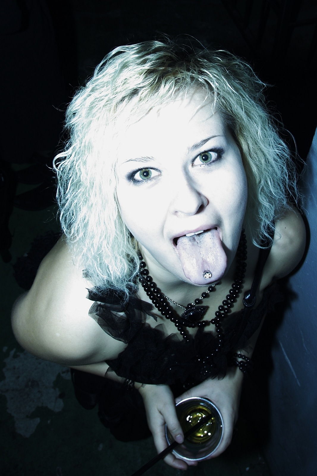 A person with light curly hair and dark eye makeup is sticking out their tongue, which has a piercing. They are holding a drink with a straw and wearing a black outfit with a necklace made of beads. The high-contrast lighting gives the Halloween party image an edgy feel.