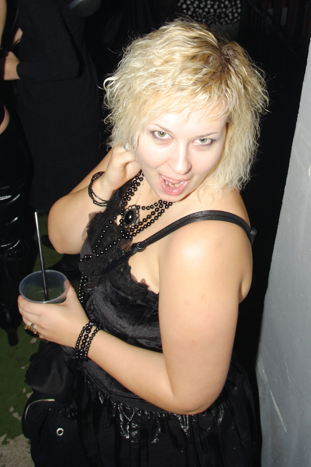 A woman with curly, platinum blonde hair is posing indoors. She is wearing a sleeveless black dress, layered black beaded necklaces, and bracelets. Holding a drink with a straw in her left hand, she gazes at the camera with an open-mouthed, playful expression reminiscent of Elektra '09 at a Halloween party.
