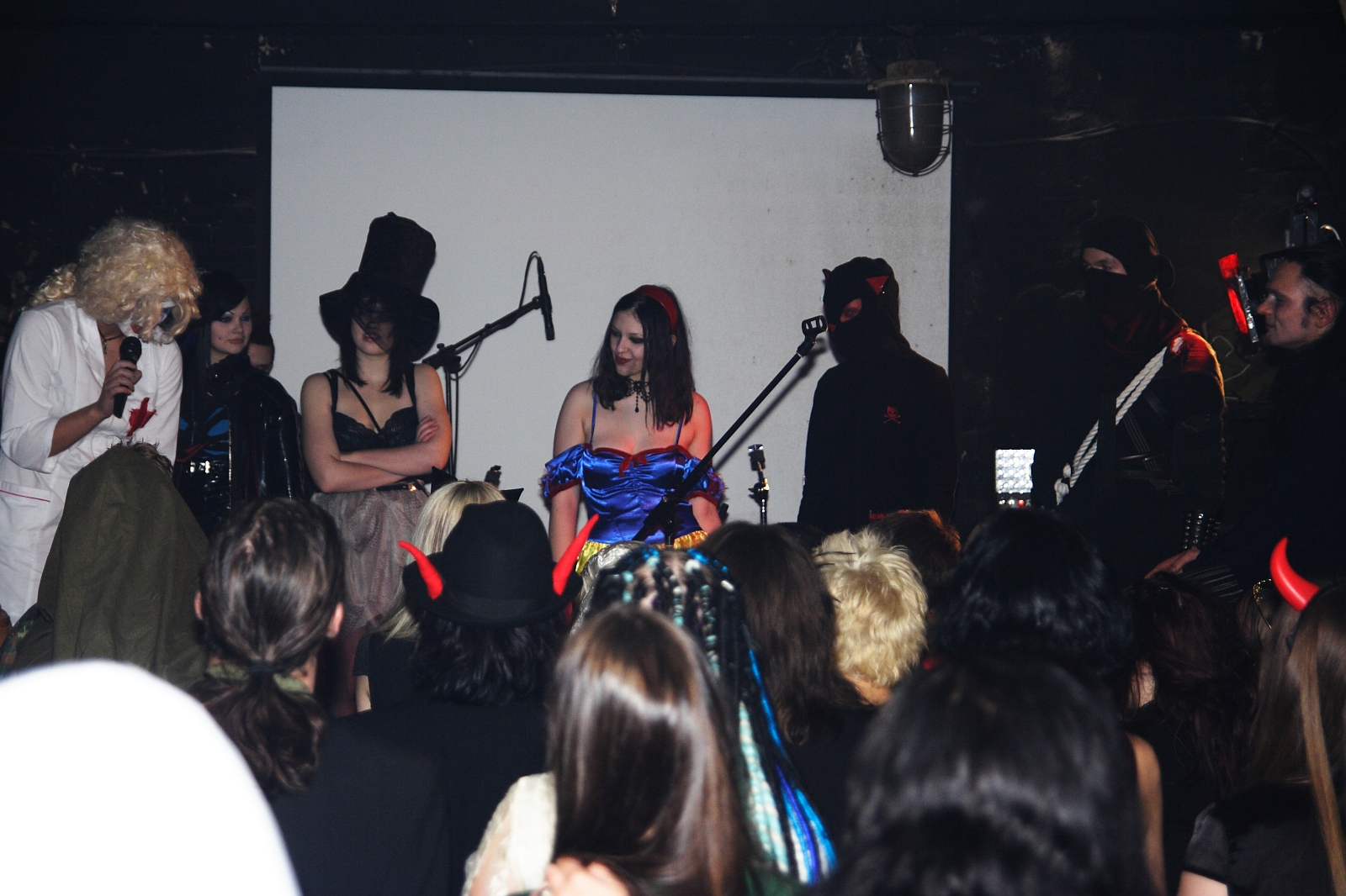 At an Elektra '09 Halloween party, a group of people dressed in various gothic and dark-themed costumes stand on stage in front of a microphone, with an audience watching them. The costumes include a white hooded cloak, tall black hats, a blue dress, black masks, and devil horns. The background is a dimly lit room with a white screen.