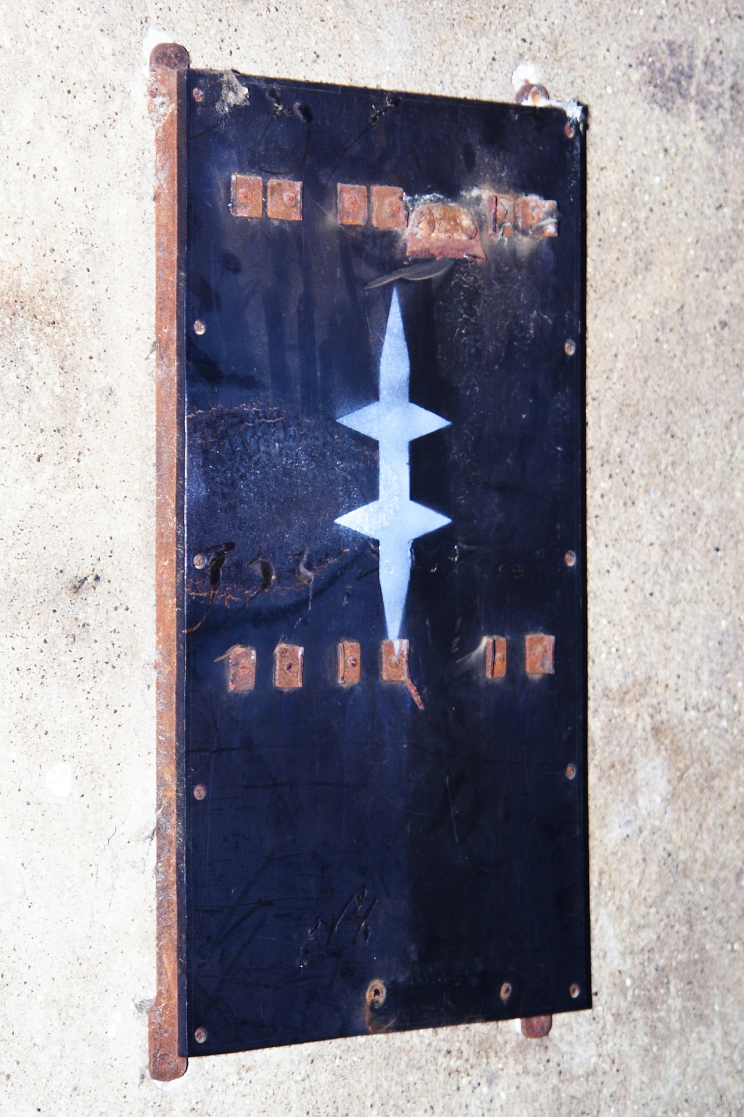 A vertical rectangular metal panel with rusted edges is affixed to a rough concrete wall. The panel features a large white abstract design resembling a stylized, symmetrical double-arrow in the center. Small copper-colored elements, reminiscent of Elektra '09, are visible near the top and bottom edges of the panel.