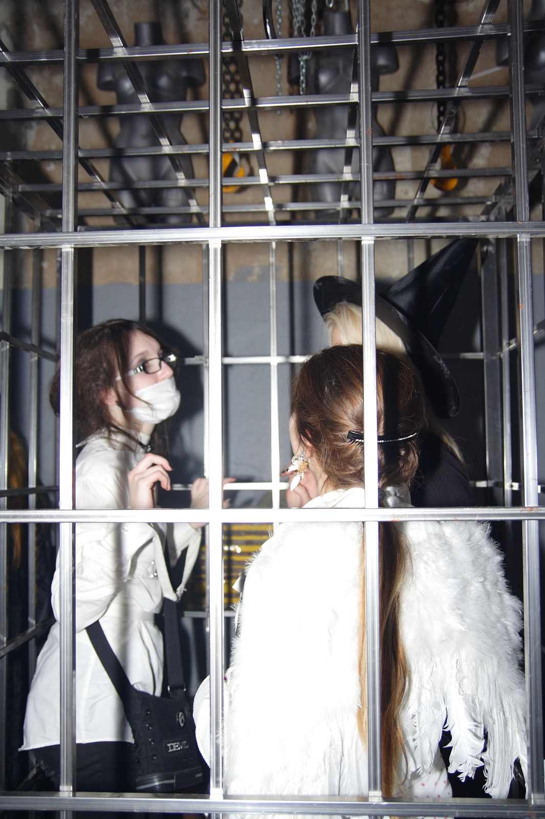 Three people are standing inside a metal cage at the Halloween party. The person on the left wears glasses, a white outfit, a mask, and carries a black bag. The person in the center, with long hair, is seen from the back wearing a white feathered garment reminiscent of Elektra '09. The person on the right wears a tall witch's hat.
