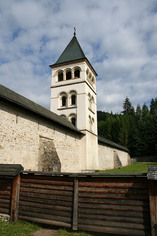 An imposing white bell tower with a conical roof stands adjacent to a solid stone church in Romania, flanked by a verdant forest under a blue sky with scattered clouds, enclosed by a rustic wooden