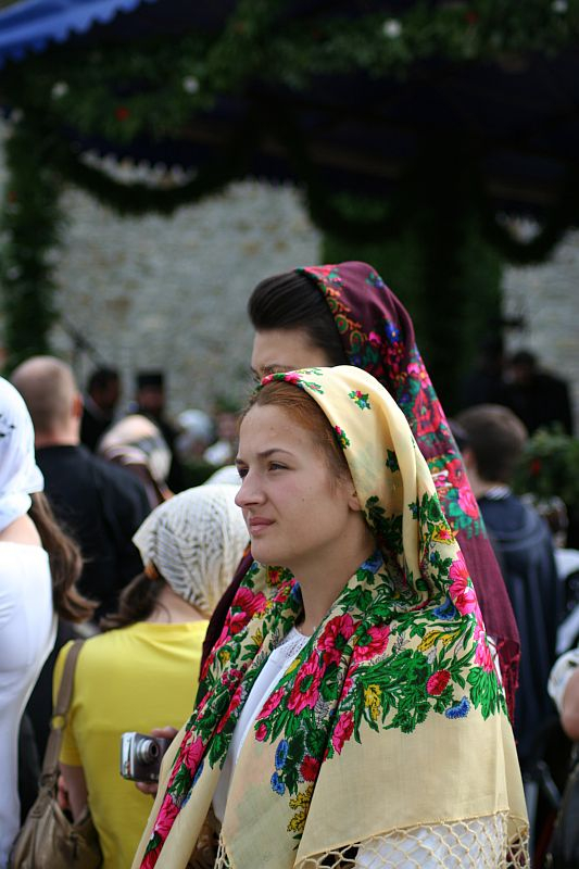 A woman in a vibrant floral headscarf attends a cultural or religious gathering in Romania, her expression contemplative, amidst a crowd of similarly dressed individuals.