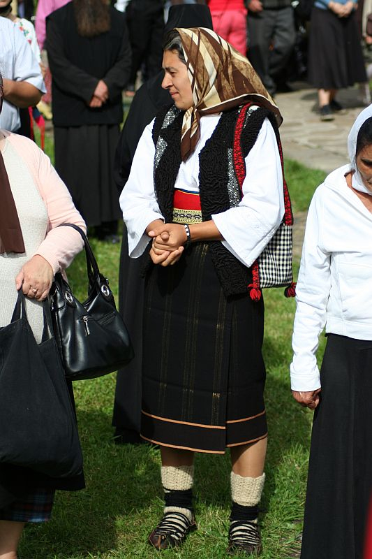 A woman, clad in traditional Romanian attire with a patterned headscarf, embroidered vest, and striped skirt, stands amidst a gathering, her hands clasped together, showcasing cultural heritage and attire.