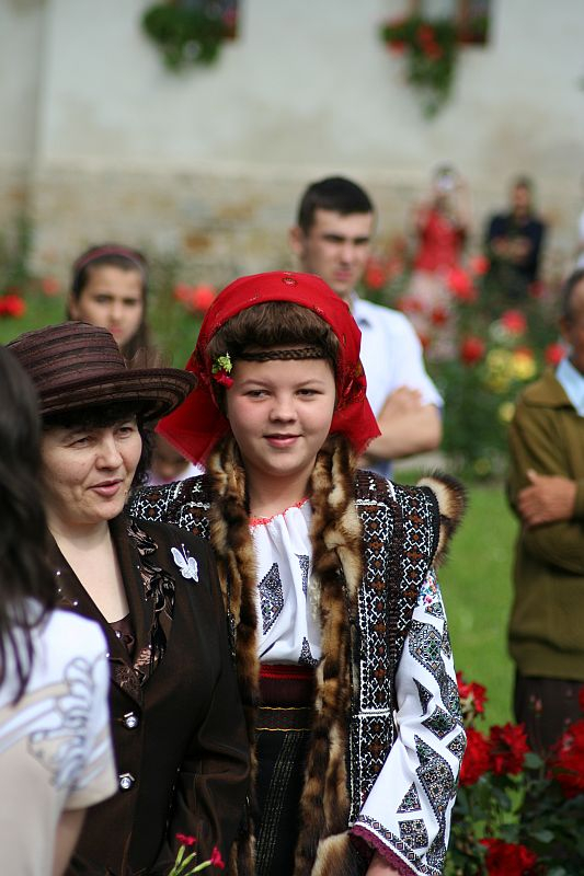 A young individual in traditional Romanian embroidered attire, including a red headscarf, confidently walks alongside a woman wearing a brown hat, as others gather amidst a vibrant floral backdrop, hinting at a cultural