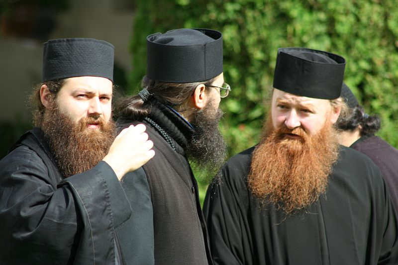 Three orthodox christian clergy members from Romania in traditional black vestments and caps engage in a conversation, with two of them facing the camera showcasing their distinct beards while the third, with his back turned,