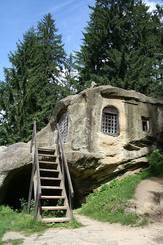 A rustic wooden staircase leads up to an old, stone hermitage built into a large boulder in Romania, surrounded by lush greenery and tall trees under a clear blue sky.