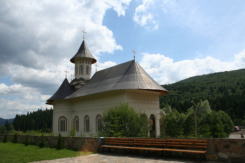 A picturesque church with a prominent bell tower and shingled roof, nestled in a serene green landscape of Romania, with fluffy clouds above and a welcoming bench at the forefront.