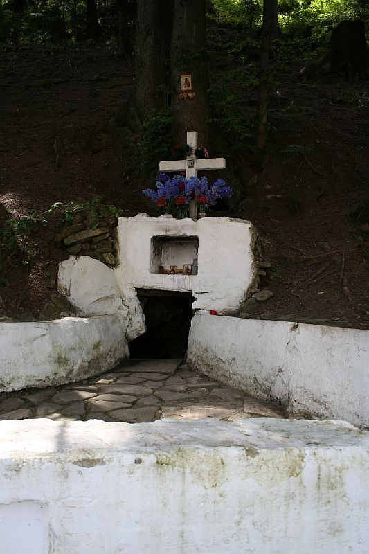 A secluded forest shrine in Romania with a white fountain structure, adorned with crosses and vibrant blue flowers, offering a tranquil spot for reflection amidst the trees.
