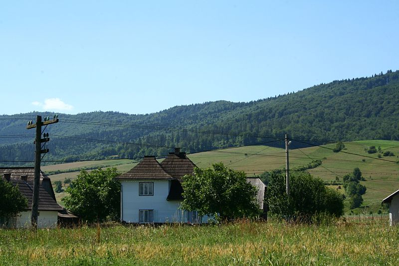 Rural landscape in Romania with traditional houses against a backdrop of rolling, forest-covered hills under a clear blue sky.