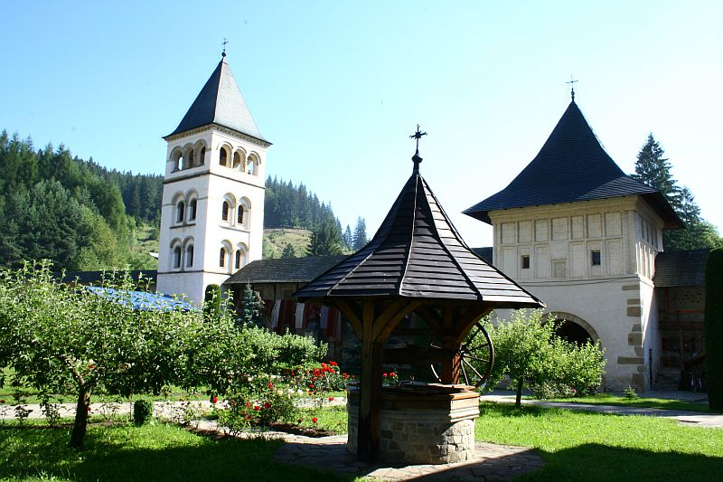 A serene monastery garden in Romania with a wooden gazebo, surrounded by lush trees and flowers, flanked by the monastery's bell tower and gatehouse on a bright sunny day.