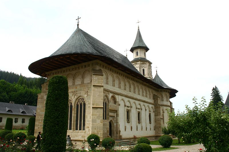 An old, ornate stone church with a conical roof and a tall bell tower, surrounded by manicured hedges and blooming flowers, stands against a cloudy sky in Romania.