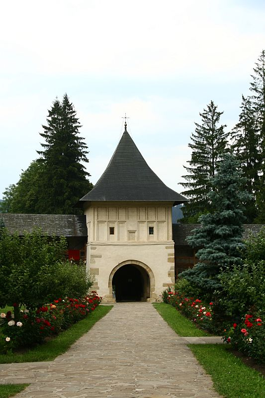 A tranquil pathway leads to a traditional stone gatehouse with a pointed roof, surrounded by lush greenery and vibrant red roses, evoking a serene, historical ambiance in Romania.