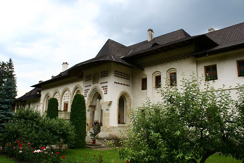 An elegant traditional monastery in Romania with an ornate porch, surrounded by a manicured lawn and blooming roses under a cloudy sky.