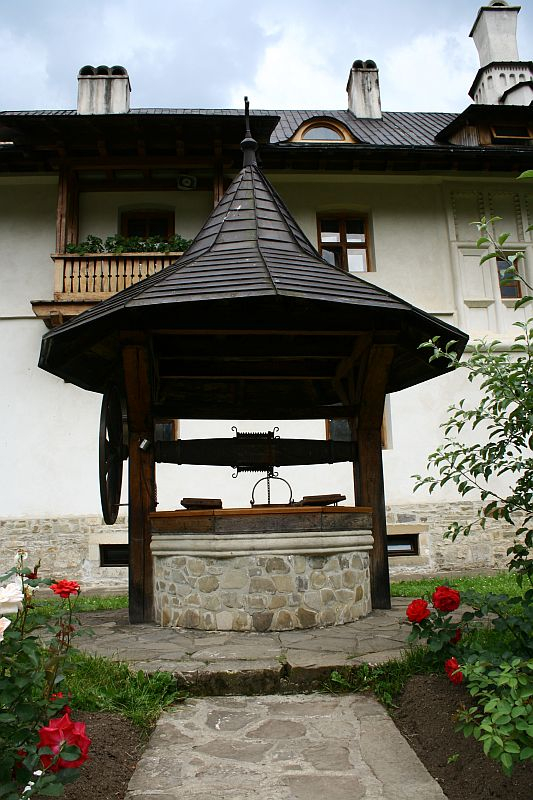 A traditional wooden well with a shingled roof, featuring a wrought iron crank, stands in the courtyard of an elegant Romania-style house with decorative eaves, framed by blooming red roses.