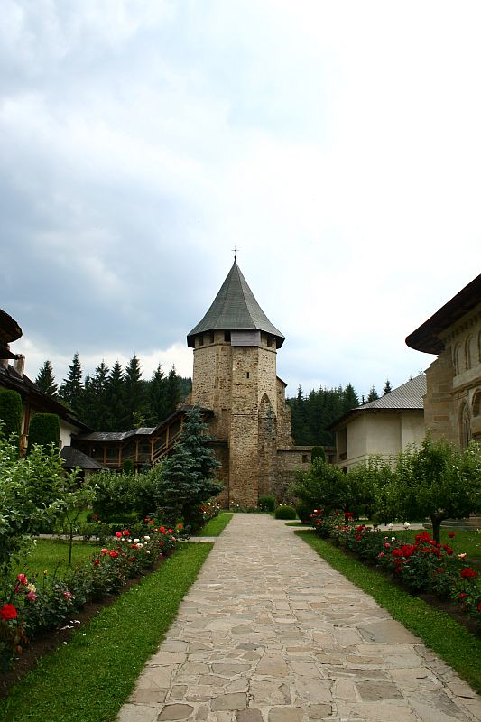A serene pathway lined with blooming flowers leads to an ancient stone tower in Romania under a cloudy sky, suggestive of a rich history nestled within a peaceful garden.
