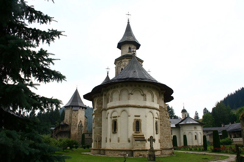 A historic church with a characteristic spire, clad in white, stands amidst greenery, with an old fortification tower visible in the background, illustrating an ancient Romanian architectural ensemble in a tranquil, forest
