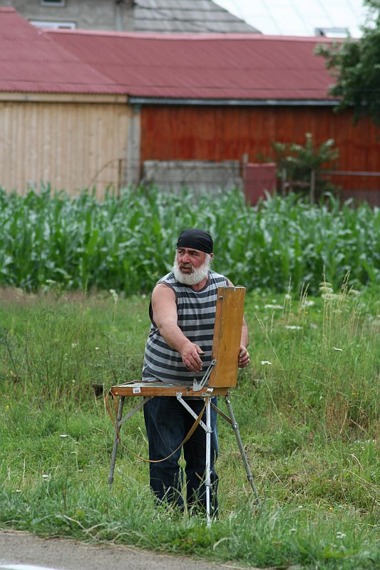 An artist in a striped tank top and black headband stands by an easel, painting en plein air amidst rural Romanian scenery with greenery and a red-roofed building in the backdrop.