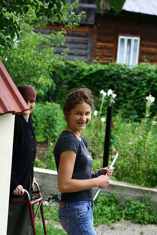 A smiling young woman in casual attire turns towards the camera while an older person leans against a wall in the background, with a quaint wooden cabin and lush greenery setting a serene backdrop in Romania.